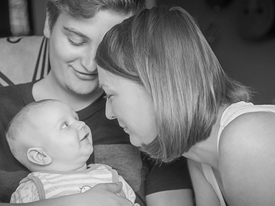 Ein junges Paar schaut liebevoll auf ihr Baby, das lächelnd zu seiner Mutter aufblickt. Die Schwarz-Weiß-Aufnahme fängt die intime und glückliche Familiendynamik in einem Heimstudio-Setting ein. Ein berührendes Familienporträt von Michael Schalansky.